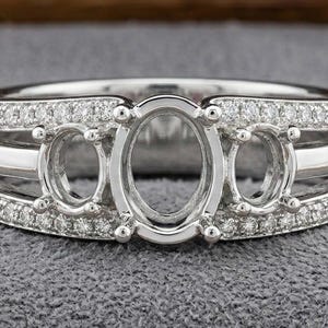 Puede incluir: Un anillo plateado con tres engastes ovalados, flanqueados por engastes redondos más pequeños. El anillo está incrustado con pequeñas piedras preciosas transparentes a lo largo de las bandas superior e inferior, creando un efecto brillante. El anillo está sobre un fondo gris.