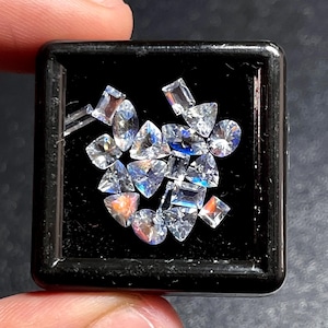 Puede incluir: Una colección de gemas facetadas de varias formas, incluyendo triángulos, cuadrados y óvalos, exhibidas en un contenedor cuadrado negro. Las gemas exhiben una gama de colores y brillan, reflejando la luz. Las gemas son probablemente para hacer joyas.