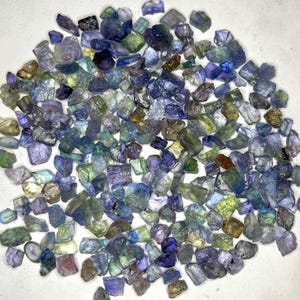 May include: A collection of raw, uncut gemstones in various shades of blue, green, and purple. The gemstones are scattered, showcasing their natural, irregular shapes and textures. The image highlights the diverse colors and translucence of the stones.