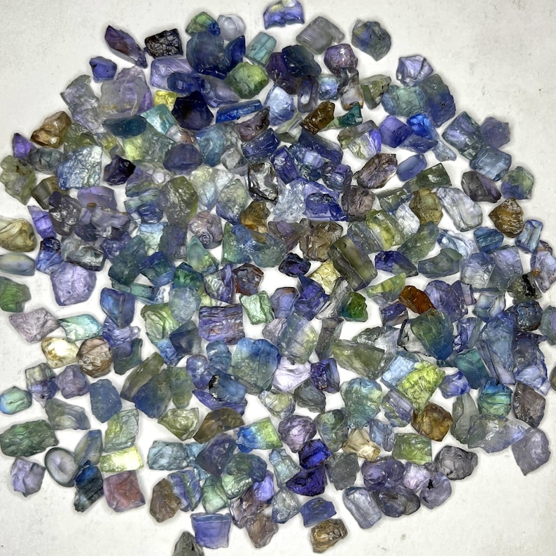 May include: A collection of raw, uncut gemstones in various shades of blue, green, and purple. The gemstones are scattered, showcasing their natural, irregular shapes and textures. The image highlights the diverse colors and translucence of the stones.