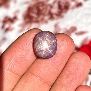 Könnte beinhalten: Ein polierter, oval geformter Edelstein mit einem gräulich-violetten Farbton. Der Stein hat ein sternförmiges Muster auf seiner Oberfläche mit einer schimmernden, reflektierenden Qualität. Der Edelstein wird in einer Hand gehalten, um seine Größe und Details zu zeigen.