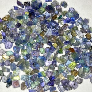 May include: A collection of raw, uncut gemstones in various shades of blue, green, and purple. The gemstones are irregularly shaped and appear to be scattered across a white surface. The stones vary in size and translucency, with some appearing more opaque than others.