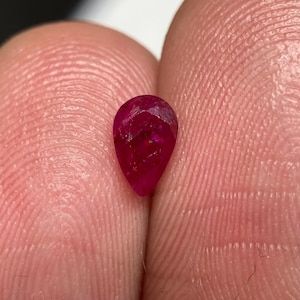 May include: A pear-shaped, deep red ruby gemstone with a faceted surface. The stone displays a translucent quality with visible internal inclusions and natural variations in color intensity throughout its structure, reflecting light across its cut facets.