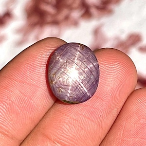 May include: A round, cabochon-cut star ruby gemstone featuring a translucent, purplish-pink hue. The stone displays a distinct six-rayed star effect, known as asterism, created by light reflecting off internal needle-like inclusions within the gem.