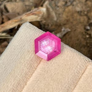 May include: A pink hexagonal gemstone with a faceted cut. The gemstone is set on a white velvet surface.