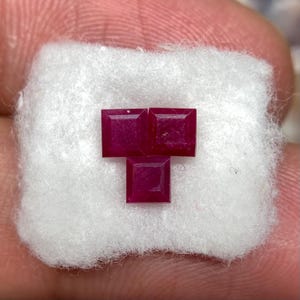 Puede incluir: Tres rubíes de color rojo intenso y corte cuadrado dispuestos en forma de T. Las piedras preciosas están facetadas con bordes afilados y una superficie pulida, descansando sobre un pequeño trozo rectangular de algodón blanco fibroso.