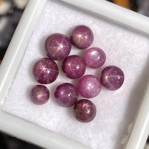 Puede incluir: Una colección de diez gemas redondas y pulidas, cada una mostrando un efecto de estrella. Las gemas son de un color rojo-púrpura intenso, con un patrón sutil en forma de estrella visible en sus superficies. Se presentan en un contenedor blanco cuadrado.
