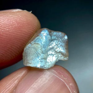 Könnte beinhalten: Ein kleiner, roher, durchscheinender Edelstein mit unregelmäßiger, gezackter Form. Der Stein zeigt eine schimmernde, irisierende Oberfläche mit blassblauen und silbernen Reflexen, die das Licht auf seinen texturierten, unebenen Facetten bricht.