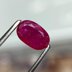 May include: A vibrant, oval-cut ruby gemstone with a deep magenta-red hue and multifaceted surface. The gemstone is held by a pair of silver-toned metal tweezers, showcasing its translucent quality and internal inclusions under bright lighting.