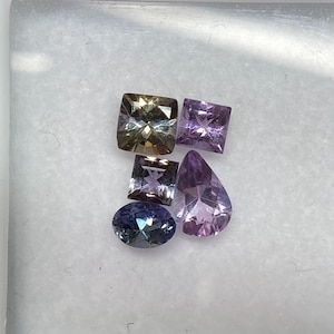 May include: A collection of five loose faceted gemstones including a square cushion-cut brownish-gold stone, a square purple stone, a smaller square purple stone, a pear-shaped light purple stone, and an oval blue-purple stone with facets.