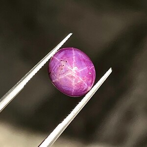 May include: A vibrant, oval-shaped magenta star ruby gemstone featuring a distinct six-rayed star pattern across its polished, cabochon-cut surface. The gemstone is held securely by a pair of silver-toned metal tweezers, showcasing its deep colour.