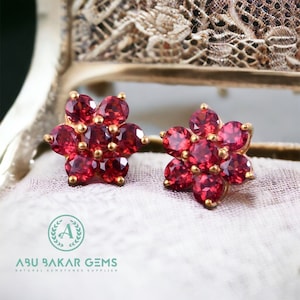 May include: A pair of gold-toned flower-shaped stud earrings. Each earring features seven oval-cut, deep red gemstones arranged to resemble a flower. The earrings are set against a soft, neutral background.