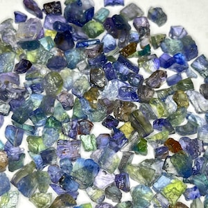 May include: A collection of raw, uncut gemstones in various shades of blue, green, and purple. The stones are irregularly shaped and appear to be tumbled or naturally formed. The image is a close-up, showcasing the texture and color variations of the gemstones.