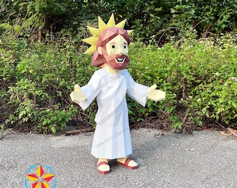 May include: A papercraft figure of Jesus with a geometric design. The figure is wearing a white robe, red sandals, and a yellow halo. The figure has a brown beard and hair. The figure is standing on a gray surface with green foliage in the background.
