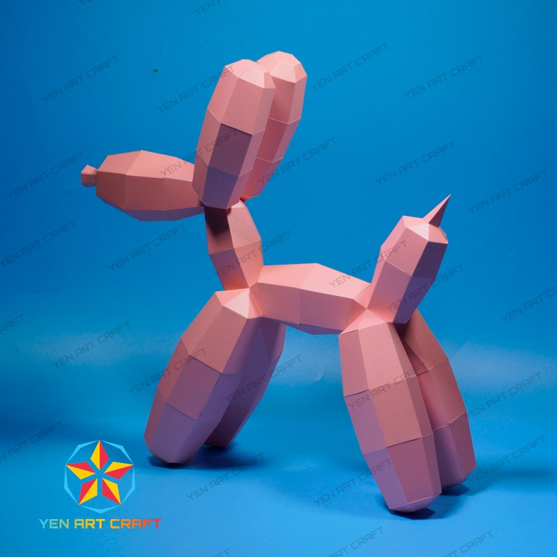 May include: A pink paper sculpture of a balloon dog, set against a blue background. The dog is constructed from geometric shapes, with visible seams and edges. The sculpture has a pointed tail and ears. The logo "YEN ART CRAFT" is visible.