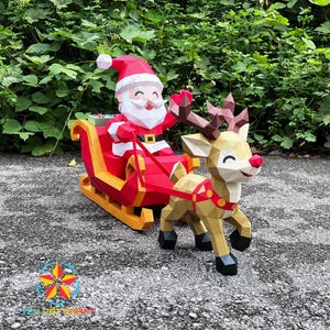 May include: A low-poly papercraft model of Santa Claus sitting in a red and yellow sleigh pulled by a tan reindeer with brown antlers. The figures feature geometric facets, a white beard, a red hat, and a red harness connecting the sleigh to the deer.