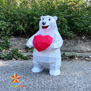 May include: A white geometric polar bear sculpture holding a red heart. The bear has a smiling expression and is made of folded paper or cardstock. The sculpture is standing on a paved surface with greenery in the background.
