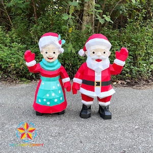 May include: Two festive papercraft figures of Santa Claus and Mrs. Claus. Santa is in a red suit with a black belt and boots. Mrs. Claus wears a red dress with a turquoise apron. Both are waving, with white beards and hair, perfect for Christmas decorations.