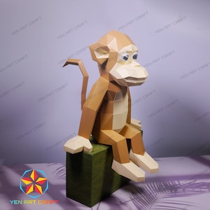 Könnte beinhalten: Ein Papiermodell eines Affen, der auf einem grünen Würfel sitzt. Der Affe ist aus braunem und beigefarbenem Papier gefertigt und hat einen langen Schwanz.