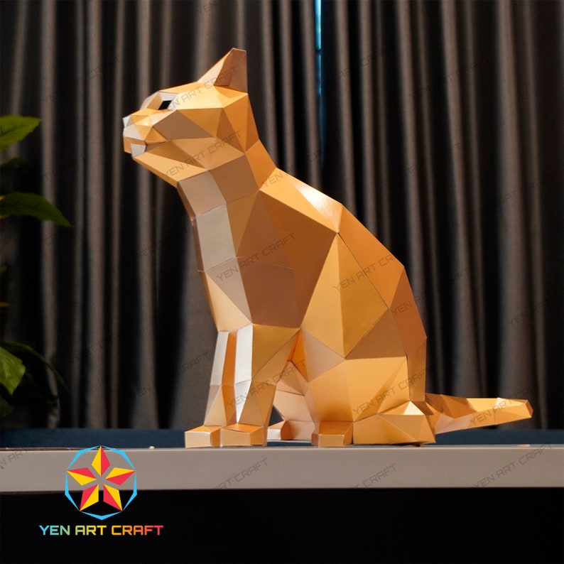 Pu&ograve; includere: Un modello di carta di un gatto color oro seduto con la coda arrotolata dietro di lui. Il modello &egrave; composto da molti piccoli triangoli e ha un aspetto geometrico. Il gatto &egrave; seduto su una superficie bianca.