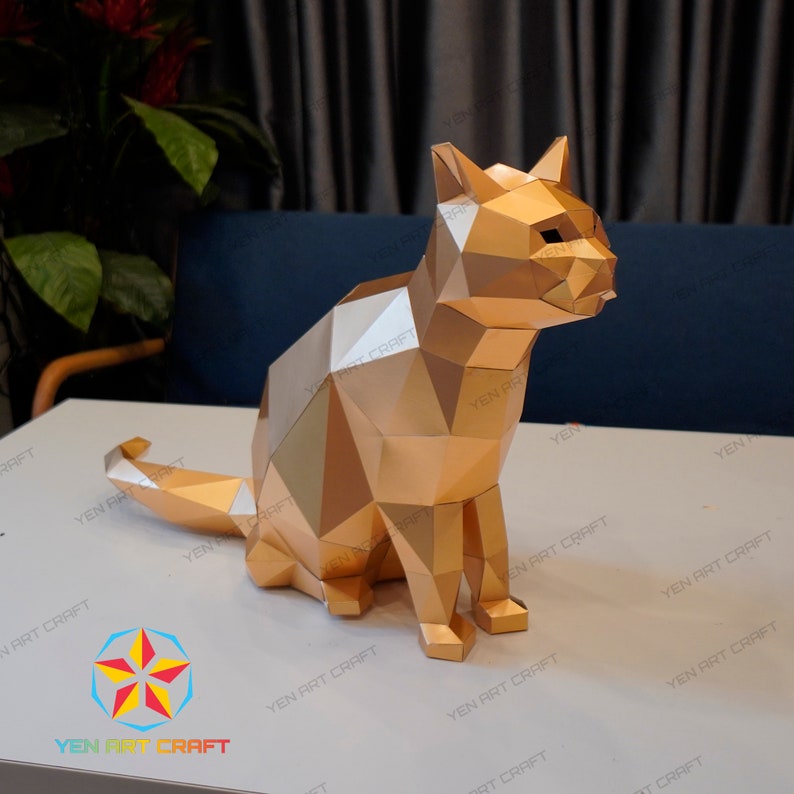 Pu&ograve; includere: Un modello di carta di un gatto dorato seduto con la coda arrotolata intorno alle zampe. Il gatto &egrave; fatto di molti piccoli triangoli che creano uno schema geometrico.