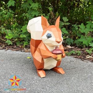 Peut inclure: Une sculpture géométrique en papier d'un écureuil, dans des tons de marron, blanc et beige, tenant un petit gland. La sculpture a un design polygonal et est placée sur un fond naturel. Le texte "YEN ART CRAFT" est visible.