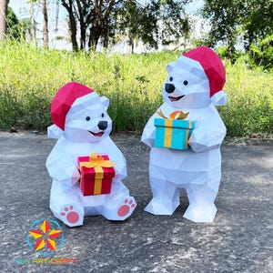 May include: Two white papercraft polar bears wearing red Santa hats, holding colorful gift boxes. The bears are standing on a paved surface with green grass and trees in the background. The text "YEN ART CRAFT" is visible.
