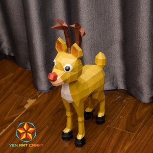May include: A papercraft reindeer model with a red nose, brown antlers, and a yellow and white body. The reindeer is standing on a wooden surface.