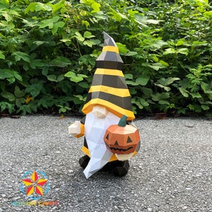 May include: A papercraft Halloween gnome with a black and yellow striped hat and a white beard. The gnome is holding an orange pumpkin with a carved face. The gnome is standing on a gray surface with green foliage in the background.