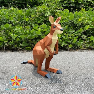 May include: A papercraft kangaroo sculpture in shades of brown, tan, and grey. The kangaroo is standing on a grey surface with green foliage in the background. The sculpture has a detailed, polygonal design.