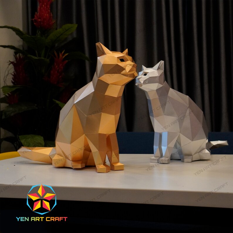 Pu&ograve; includere: Due gatti di carta origami, uno dorato e uno argentato, sono seduti uno accanto all'altro su una superficie bianca. I gatti si guardano e sembrano baciarsi. L'immagine include il testo "YEN ART CRAFT".