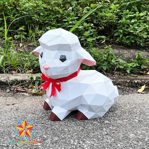 Peut inclure: Un agneau en papier blanc géométrique avec un nœud rouge autour du cou. L'agneau a des oreilles intérieures roses et des sabots bruns. Le texte "YEN ART CRAFT" est visible.