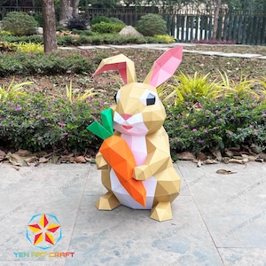 Peut inclure: Une sculpture géométrique en papier représentant un lapin beige, rose et orange, tenant une grande carotte. Le lapin a de grandes oreilles et est posé sur une surface pavée avec de la verdure en arrière-plan. L'œuvre est étiquetée "YEN ART CRAFT".
