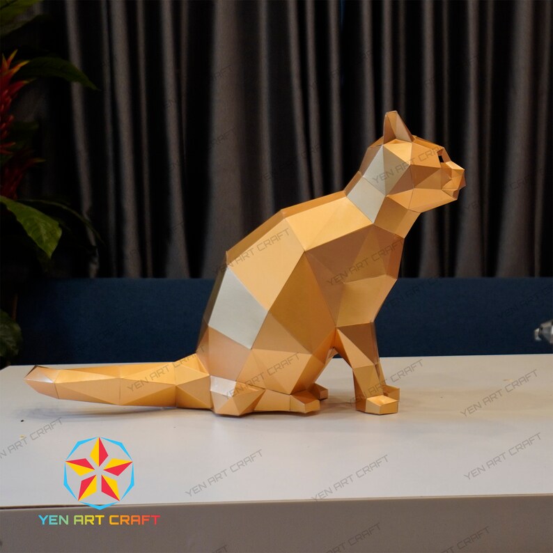 Pu&ograve; includere: Una scultura di gatto origami dorata con una striscia bianca sul petto. La scultura &egrave; seduta su una superficie bianca.
