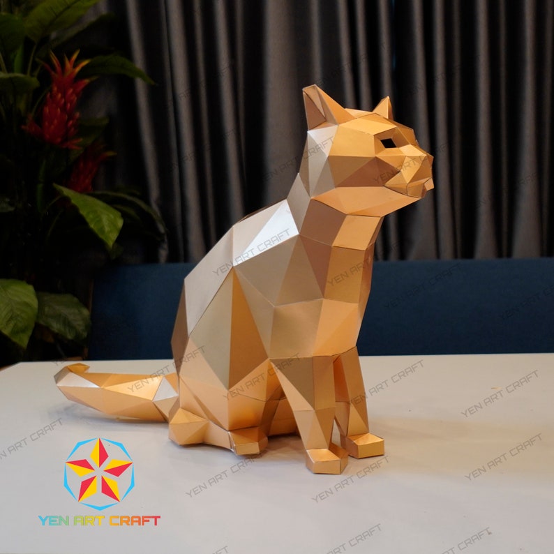 Pu&ograve; includere: Un modello di carta di un gatto dorato seduto con la coda arrotolata intorno alle zampe. Il modello &egrave; fatto di molti piccoli triangoli che creano uno schema geometrico.
