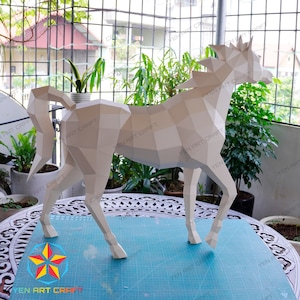 May include: A white papercraft horse model with a geometric design. The horse is standing on a blue cutting mat with a white grid pattern. The model is made of folded paper and has a three-dimensional appearance.