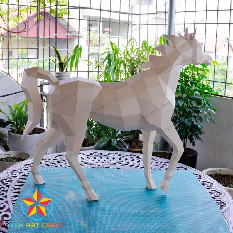May include: A white papercraft horse model with a geometric design. The horse is standing on a blue cutting mat with a white grid pattern. The model is made of folded paper and has a three-dimensional appearance.