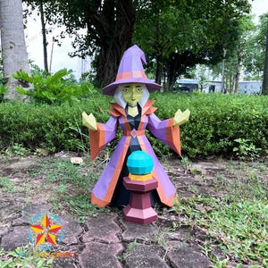 Peut inclure: Une figurine en papier low-poly représentant une sorcière vêtue d'un chapeau pointu et d'une robe violette et orange, avec des cheveux blancs et un visage vert. La sorcière se tient derrière un piédestal rouge foncé surmonté d'une sphère.