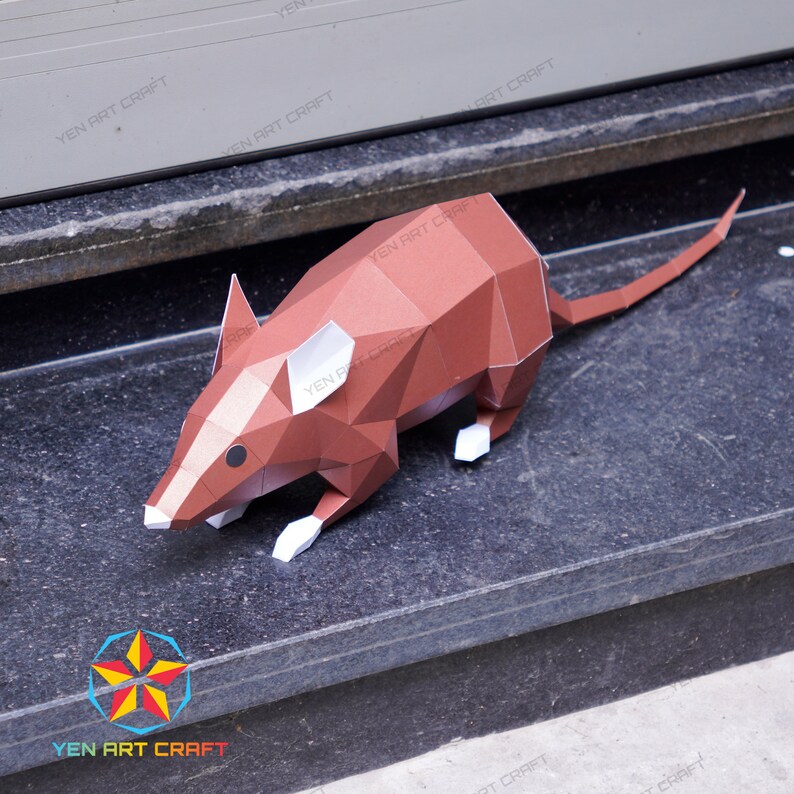 May include: A brown papercraft mouse model with white accents. The mouse is posed in a walking position with its tail extended behind it. The model is sitting on a gray stone surface.