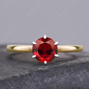 May include: A gold engagement ring with a red gemstone set in a six-prong setting.