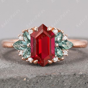 May include: A rose gold ring featuring a large, faceted, rectangular ruby center stone. The ruby is surrounded by smaller, light green gemstones and small white diamonds. The ring is set on a gray surface.