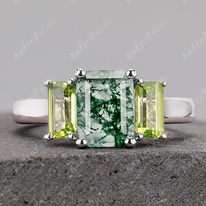 May include: A silver ring featuring a green and white emerald cut gemstone flanked by two smaller, square-shaped, light green gemstones.