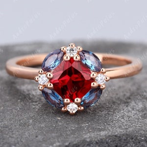 May include: A rose gold engagement ring featuring a square-cut red gemstone surrounded by smaller white gemstones and blue gemstones.