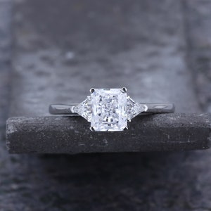 Pode incluir: Um anel de noivado de prata com um grande diamante retangular transparente no centro e dois diamantes triangulares transparentes menores de cada lado.