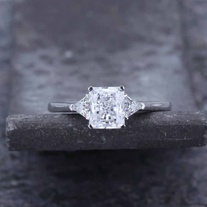 Pode incluir: Um anel de noivado de prata com um grande diamante retangular transparente no centro e dois diamantes triangulares transparentes menores de cada lado.