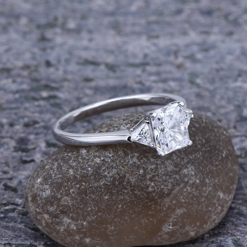 Pode incluir: Um anel de noivado de prata com um grande diamante central quadrado e dois diamantes triangulares menores de cada lado.
