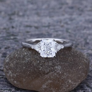 Pode incluir: Um anel de noivado de prata com um grande diamante quadrado no centro e dois diamantes triangulares menores de cada lado.