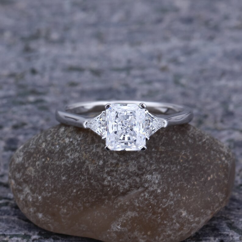 Pode incluir: Um anel de noivado de prata com um grande diamante quadrado no centro e dois diamantes triangulares menores de cada lado.