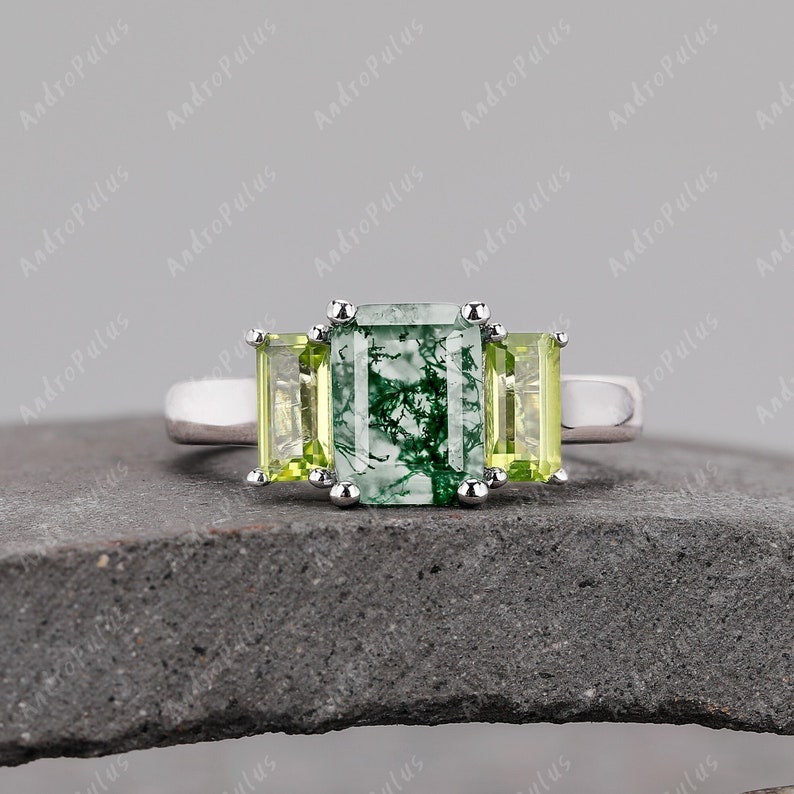 May include: A silver ring featuring a green and white emerald cut gemstone flanked by two smaller, square-shaped, light green gemstones.