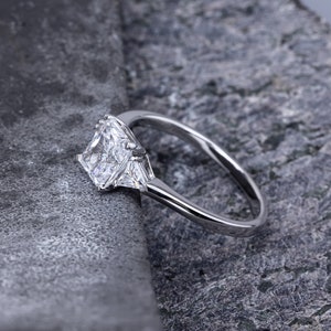 Pode incluir: Um anel de prata com um grande diamante quadrado e dois diamantes triangulares menores de cada lado.